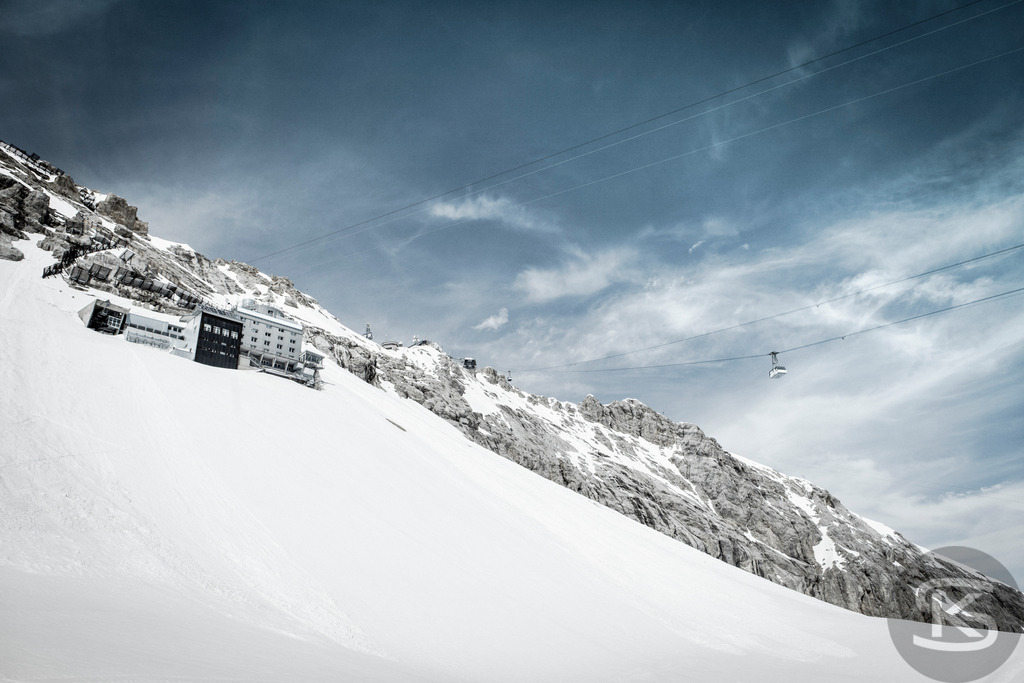 Winterlandschaft Zugspitze: Umweltforschungsstation Schneefernerhaus und Gletscherbahn | Die historische Umweltforschungsstation Schneefernerhaus auf der Zugspitze, ehemals ein mondänes Hotel und Endbahnhof der Zahnradbahn, im tiefen Winterschnee. Im Vordergrund dominiert eine steile, schneebedeckte Piste, während eine Kabine der Zugspitz-Gletscherbahn in der Ferne den Hang hinauffährt. - Realisiert mit Pictrs.com