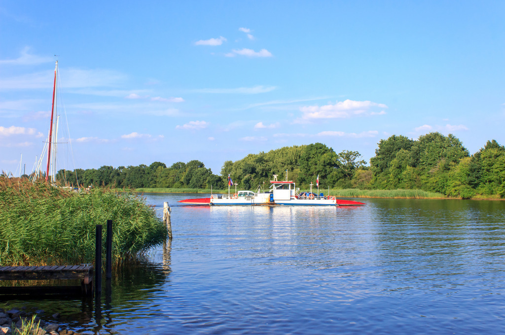 Wandbild: Schleifähre Missunde | Dieses Wandbild im Querformat zeigt die Schleifähre in Missunde im Sommer. Auf der Schlei ist eine schöne Spiegelung zu sehen. Der blaue Himmel zeigt nur wenige Wolken.  - Realisiert mit Pictrs.com