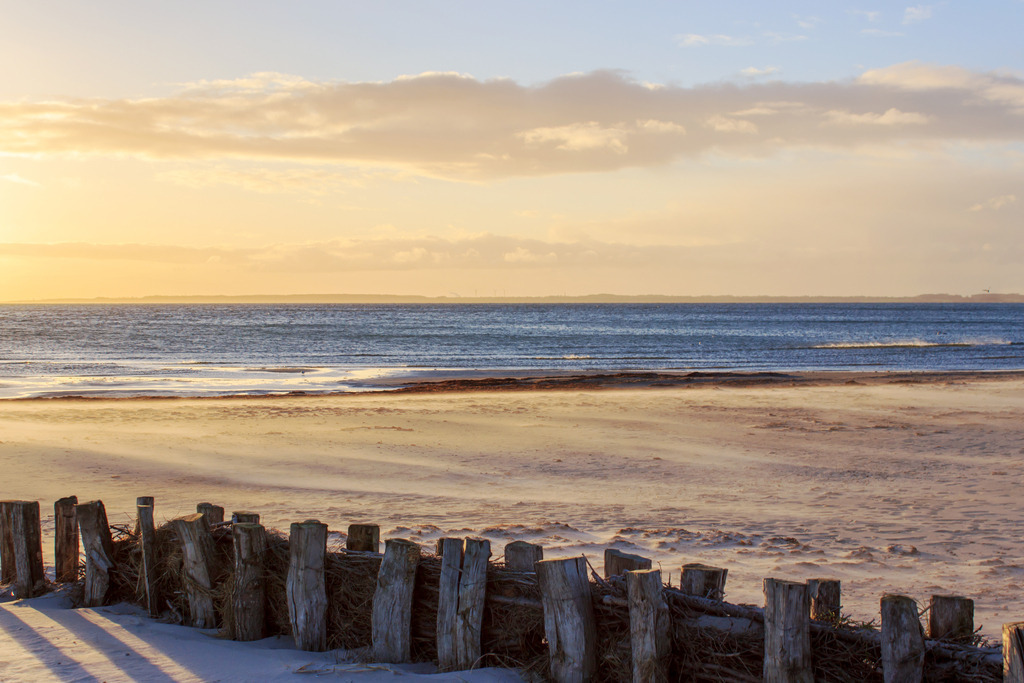 Wandbild: Morgenrot am Sandstrand | Dieses Wandbild im Querformat zeigt eine schöne Morgenstimmung am Sandstrand. Im Vordergrund ist ein Sandfang zu sehen, durch den das Licht der aufgehenden Sonne scheint. Am Himmel befinden sich schöne Wolken im Abendrot. - Realisiert mit Pictrs.com