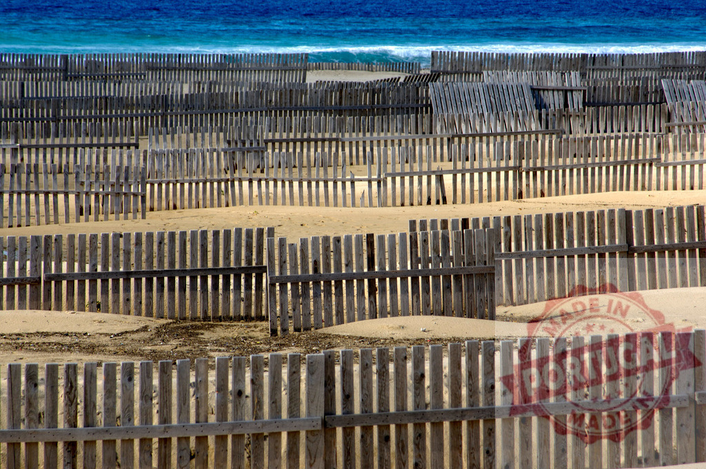 Wooden fences protecting sand dunes at the ocean. Espana - Cadiz | Wooden fences forming patterns across sand dunes at a blue ocean beach line - Realized with Pictrs.com