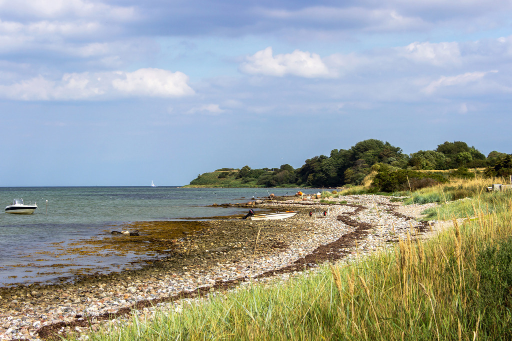 Wanbild: Ostseeidylle mit Natur und Weite in Habernis | Dieses Wandbild zeigt den ursprünglichen Naturstrand von Habernis an der Flensburger Förde – ein Ort voller Ruhe und norddeutscher Gelassenheit. Im Vordergrund liegen verstreute Steine auf dem sandigen Ufer, während sich in der Ferne die bewachsene Steilküste majestätisch erhebt. Kleine Boote schaukeln sanft auf dem Wasser, und ein Segelboot zieht gemächlich vorbei – ein Moment der Entschleunigung, eingefangen in einem Bild. Erhältlich als Leinwand, Alu-Dibond, Acrylglas, FineArt Papier oder als Akustikbild, bringt dieses Motiv die natürliche Schönheit der Ostseeküste in dein Zuhause. Ideal für Wohnräume, Büros oder Ferienwohnungen – überall dort, wo Sehnsucht nach Meer und Natur spürbar werden soll.Ein Stück Habernis für deine Wand – authentisch, beruhigend und voller maritimer Atmosphäre. - Realisiert mit Pictrs.com