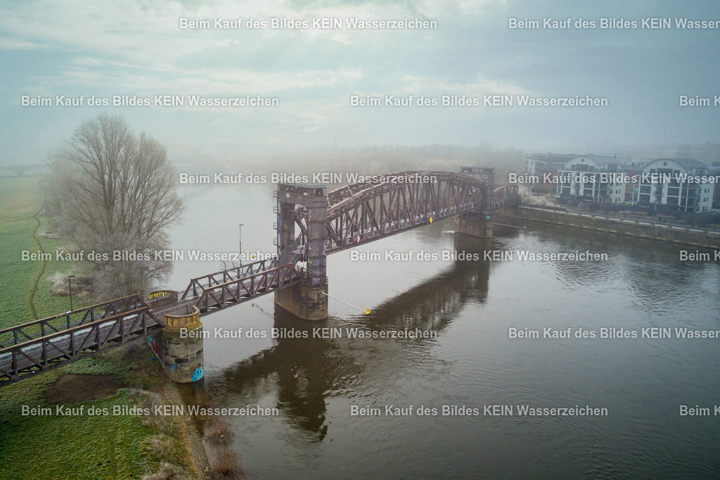 Hubbrücke_Reif_Nebel_Winter_Stadtpark-0017 | Das Luftbild zeigt die historische Hubbrücke in Magdeburg aus einer Drohnenperspektive an einem nebligen Morgen. Der leichte Nebel umhüllt die Stahlkonstruktion und hebt ihre markanten Linien besonders eindrucksvoll hervor. Trotz der stimmungsvollen Atmosphäre bleibt die außergewöhnliche Ingenieurarchitektur des Bauwerks das zentrale Motiv der Aufnahme.Die Hubbrücke wurde ursprünglich 1848 als Eisenbahnbrücke errichtet und erhielt ihre heutige Stahlkonstruktion im Jahr 1933. Mit einer Gesamtlänge von 220 Metern und einem 90 Meter langen beweglichen Brückenteil zählt sie zu den bedeutendsten technischen Denkmalen Magdeburgs. Der rund 450 Tonnen schwere Hubteil konnte mithilfe eines Gegengewichtssystems innerhalb weniger Minuten um 2,87 Meter angehoben werden, um den Schiffsverkehr auf der Elbe auch bei Hochwasser sicher passieren zu lassen.Die Drohnenaufnahme zeigt eindrucksvoll die Konstruktion aus genieteten Stahlfachwerken und die präzise Ingenieurleistung der damaligen Zeit. Der morgendliche Nebel verleiht dem historischen Bauwerk eine besondere Atmosphäre, während die technischen Details und die markante Geometrie der Hubbrücke eindrucksvoll zur Geltung kommen. - Realisiert mit Pictrs.com