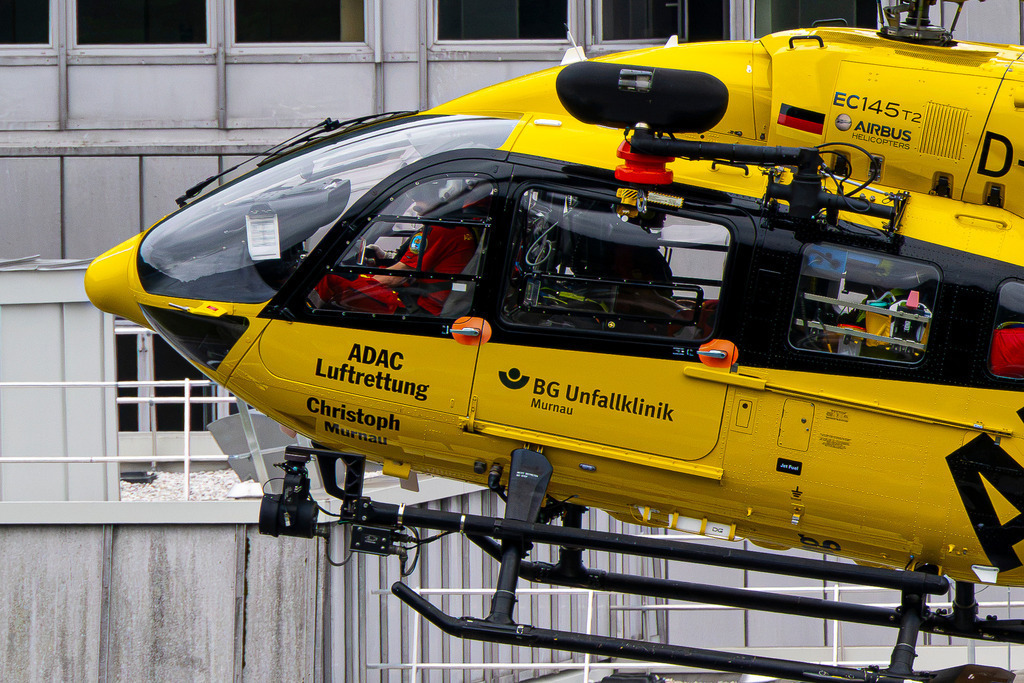Im Einsatz für das Leben: Detailaufnahme von Christoph Murnau | Eine eindrucksvolle Nahaufnahme zeigt die Cockpit-Partie des ADAC-Rettungshubschraubers „Christoph Murnau“ (EC145 T2 / Airbus Helicopters). Im Zusammenspiel mit der BG Unfallklinik Murnau symbolisiert die leuchtend gelbe Maschine höchste Präzision, Schnelligkeit und verlässliche Hilfe aus der Luft. Der Blick auf die Rettungswinde und das besetzte Cockpit vermittelt die Anspannung und Hingabe der Crew. Ein Bild, das für Hoffnung, moderne Medizin und Lebensrettung im entscheidenden Moment steht. - Realisiert mit Pictrs.com