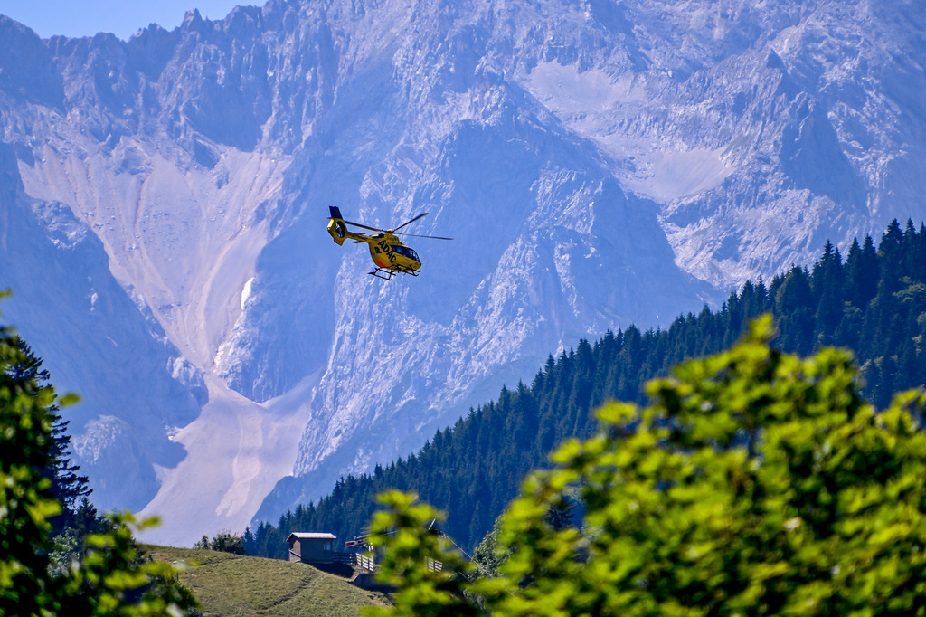 ADAC Rettungshubschrauber in den Alpen | Ein markanter gelber Rettungshubschrauber des ADAC fliegt vor der gigantischen, bläulich schimmernden Kulisse eines massiven Felsmassivs (Wettersteingebirge bei Garmisch-Partenkirchen). Im Vordergrund bilden saftig grüne Bäume einen natürlichen Rahmen, während tief unten am Hang eine kleine Holzhütte die unendliche Weite der alpinen Bergwelt unterstreicht. - Realisiert mit Pictrs.com