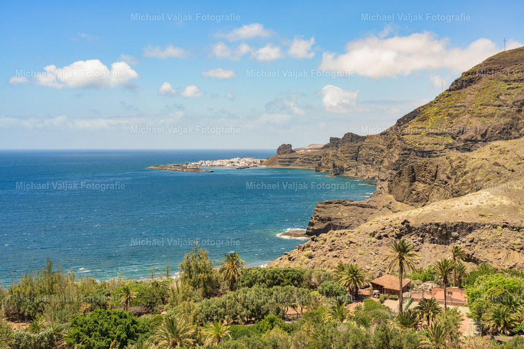 Blick nach Puerto de las Nieves auf Gran Canaria | Der Blick vom Mirador de Guayedra hinunter auf Puerto de las Nieves offenbart die wilde Schönheit der kanarischen Küste. Hoch über dem Atlantik bietet dieser Aussichtspunkt eine spektakuläre Szenerie: Vulkangestein ragt schroff in die Höhe, das Meer erstreckt sich endlos in tiefem Blau, und die Küstenlandschaft vereint raue Natur mit stiller Eleganz.Das Tal von Guayedra liegt eingebettet zwischen dunklen Felsen und üppigem Grün. Der schmale Pfad durch diese Region führt zu beeindruckenden Panoramen, die besonders im Licht der Abendsonne ihre volle Wirkung entfalten. Weit draußen am Horizont erscheint Puerto de las Nieves, ein friedlicher Fischerort, dessen weiße Häuser und ruhiger Hafen eine harmonische Ergänzung zur wilden Küstenlandschaft bilden. - Realisiert mit Pictrs.com