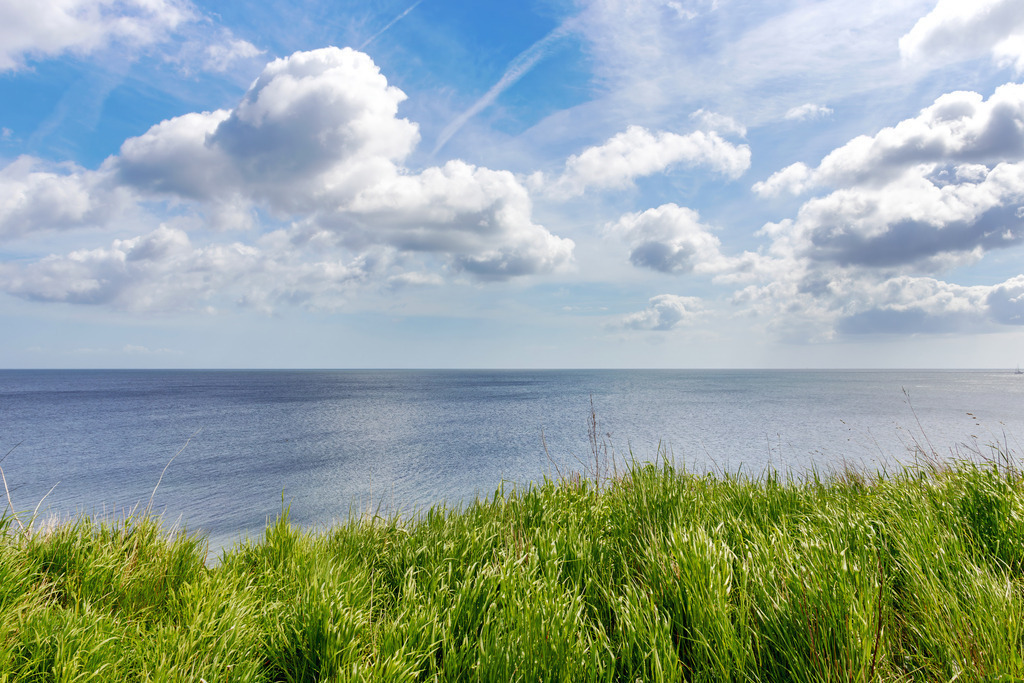 Wandbild: Blick von der Steilküste auf die Ostsee | Offene Weiten und sanfte Naturfarben sorgen für eine entspannende Atmosphäre – ideal für medizinische Einrichtungen. Dieses Wandbild zeigt die Steilküste von Schönhagen mit weitem Blick über das Meer, während das Gras im Vordergrund die natürliche Küstenlandschaft unterstreicht. Der strahlend blaue Himmel mit leichten Frühlingswolken verstärkt die beruhigende Wirkung des Motivs und sorgt für eine angenehme Raumgestaltung in Wartezimmern, Behandlungsräumen oder Empfangsbereichen. - Realisiert mit Pictrs.com