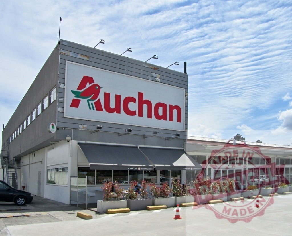Auchan hypermarket building featuring cafe frontage and parking | Auchan hypermarket building with its logo sign, adjacent cafe terrace, and parking lot - Realized with Pictrs.com