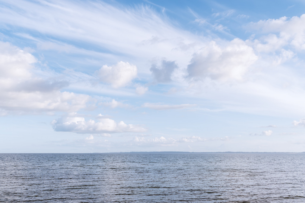 Wandbild: Weite über dem Meer – Damp | Vor Damp öffnet sich die Ostsee weit und ruhig, das Wasser liegt in sanfter Bewegung unter einem hellen Himmel mit verstreuten Wolken. Am Horizont lässt sich die gegenüberliegende Uferseite der Eckernförder Bucht erahnen, was dem Motiv eine zusätzliche räumliche Tiefe verleiht. Die klare Horizontlinie und die offene Weite schaffen eine stille, maritime Atmosphäre. Dieses Wandbild bringt die entspannte, natürliche Küstenstimmung von Damp direkt in den Raum und öffnet ihn für einen ruhigen Blick über das Meer.