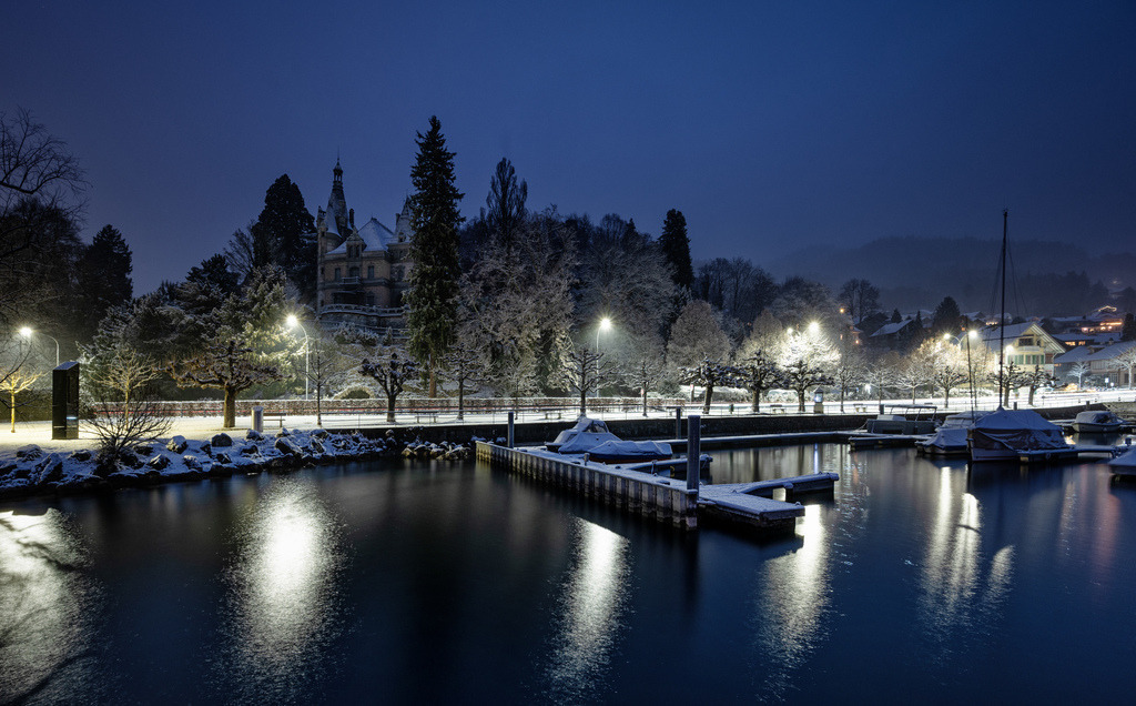 Schloss Hünegg Wintermorgen | Die winterliche Landschaft am Thunersee zeigt Schloss Hünegg und den verschneiten Hafen bei Einbruch der Dunkelheit. Strassenlaternen beleuchten die schneebedeckten Baeume und Wege, deren Licht sich ruhig im Wasser des Sees spiegelt. Die Szene vermittelt eine stille, friedliche Winterstimmung. - Realisiert mit Pictrs.com
