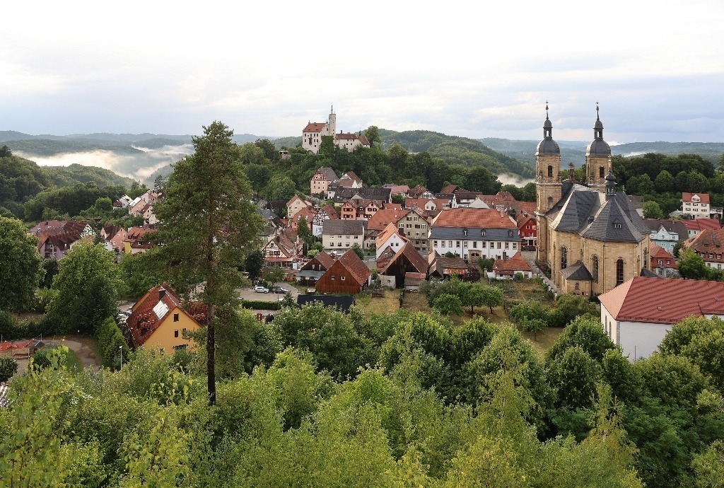 Blick vom Kreuzberg auf Gößweinstein | Das Foto zeigt den Blick vom Kreuzberg auf den Marktort Gößweinstein in der Fränkischen Schweiz. Eingebettet in bewaldete Hügel und markante Felsen liegt der Ort malerisch im Tal und wird von der weithin sichtbaren Wallfahrtsbasilika und der Burg geprägt. Die Türme der Kirche und die historischen Gebäude verleihen dem Ortsbild einen besonderen Charakter. Der aufsteigende Dunst über den Wäldern nach einem Gewitterregen geben dem Bild zusätzliche Tiefe. Das Sonnenlicht hebt die Architektur des Ortes eindrucksvoll hervor. Die Aufnahme vermittelt Ruhe und Weite und zeigt die harmonische Verbindung von Natur, Geschichte und Kultur. Der erhöhte Standort auf dem Kreuzberg eröffnet einen eindrucksvollen Panoramablick auf Gößweinstein und seine reizvolle Umgebung. - Realisiert mit Pictrs.com