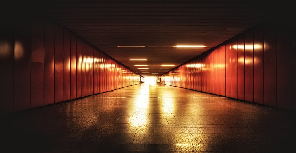Fußgängerpassage-Stuttgarter Haupbahnhof | Das Bild zeigt eine Unterführung in der Klett-Passage am Hauptbahnhof in Stuttgart. Die Passage ist bekannt für ihre markante, orangefarbene Wandverkleidung und die Beleuchtung, die eine warme Atmosphäre schafft. Sie dient als wichtiger Verkehrsknotenpunkt für Fußgänger, die zwischen dem Hauptbahnhof und der Innenstadt pendeln. Die Szene fängt die Perspektive eines langen, beleuchteten Tunnels ein. - Realisiert mit Pictrs.com