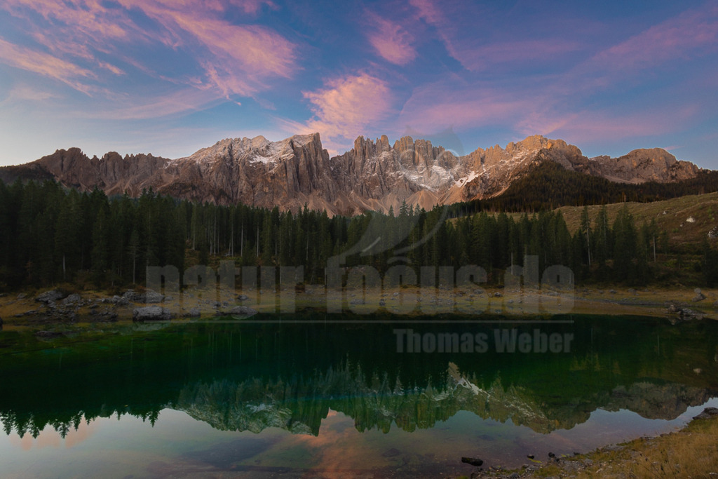 R6NF6684_20251001 | Ein atemberaubender Panoramablick auf den Karersee (Lago di Carezza) in den Dolomiten während der Dämmerung. Der Himmel ist in sanfte Rosa- und Blautöne getaucht, mit zarten Wolkenstreifen, die das Licht des Sonnenuntergangs einfangen. Im Hintergrund erheben sich majestätische, teils schneebedeckte Berggipfel der Rosengartengruppe, deren schroffe Felsformationen im warmen Licht leuchten. Ein dichter Nadelwald säumt das Ufer des Sees. Das kristallklare, smaragdgrüne Wasser des Karersees spiegelt die umliegende Berglandschaft und den farbenprächtigen Himmel perfekt wider, wodurch eine symmetrische und ruhige Szene entsteht. Es sind keine Tiere oder Interaktionen zu erkennen. - Realisiert mit Pictrs.com
