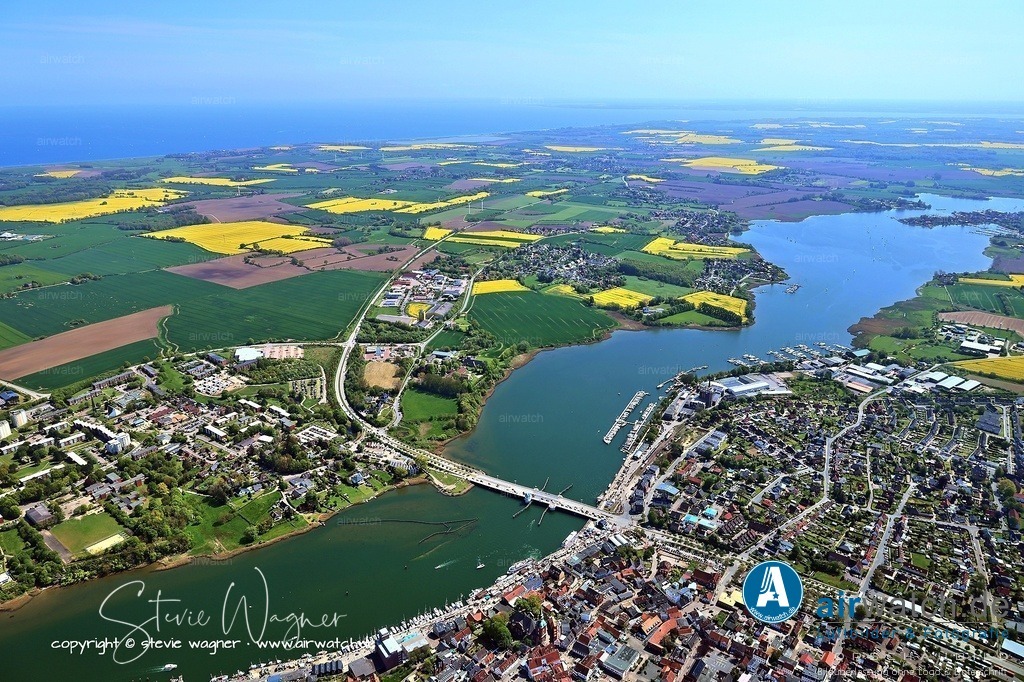 Luftbild Kappeln an der Schlei & Ellenberg | Die Geschichte der Schleibrücke in Kappeln reicht bis ins Jahr 1671 zurück, als eine Fährverbindung zwischen Kappeln und Ellenberg nachgewiesen wurde. Die erste Brücke an dieser Stelle wurde 1850 gebaut, obwohl über sie wenig bekannt ist. Im Jahr 1867 wurde auf Initiative Kappelner Kaufleute eine Pontonbrücke errichtet, die 180 Meter lang war und auf 18 Krähnen ruhte, um die tiefste Stelle der Schlei zu überqueren. Diese Brücke diente auch als Schienenführung für die Eckernförder Kreisbahn, wobei die Lokomotiven jedoch zu schwer waren und die Wagen daher mit Pferden über die Brücke gezogen werden mussten. Für die Nutzung der Brücke war Brückengeld fällig, und sie wurde durch klimatische und schiffsverkehrsbedingte Widrigkeiten regelmäßig beschädigt.