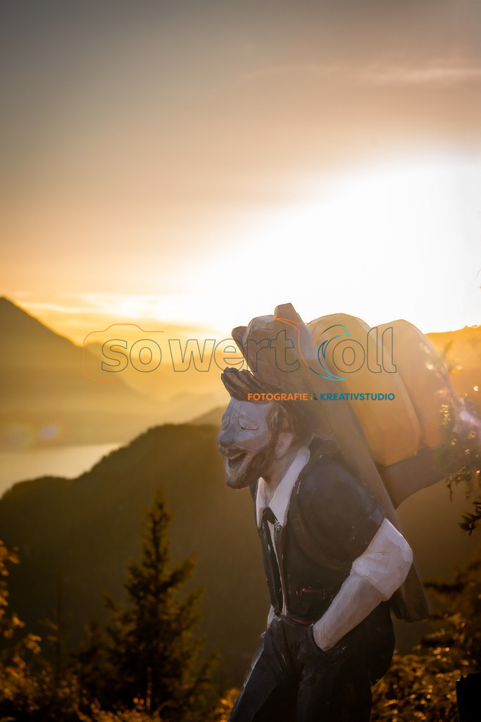 Goldenes Licht am Harder Kulm | Dieses stimmungsvolle Bild wurde während des goldenen Stundenlichts auf dem Harder Kulm aufgenommen – einem der schönsten Aussichtspunkte des Berner Oberlandes. Im Vordergrund steht eine kunstvoll geschnitzte Figur, deren fröhlicher Ausdruck die Tradition und Kultur  widerspiegelt. Im Hintergrund breiten sich die sanften Silhouetten der Alpen und der funkelnde See im warmen Abendlicht aus.Ein Zusammenspiel aus Natur, Kultur und einzigartiger Lichtstimmung – perfekt für Liebhaber stimmungsvoller Landschaftsfotografie.