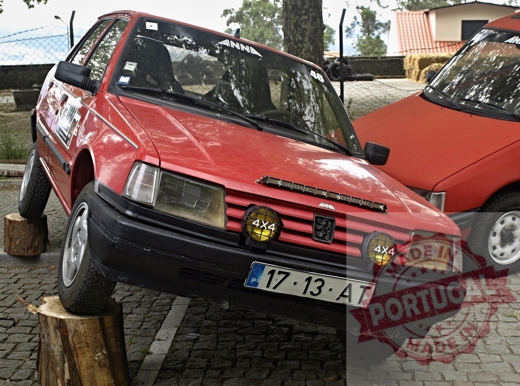 Red Peugeot hatchback balancing on tree stumps, off-road testing | Modified red Peugeot hatchback demonstrating suspension articulation on wooden tree stumps - Realisiert mit Pictrs.com