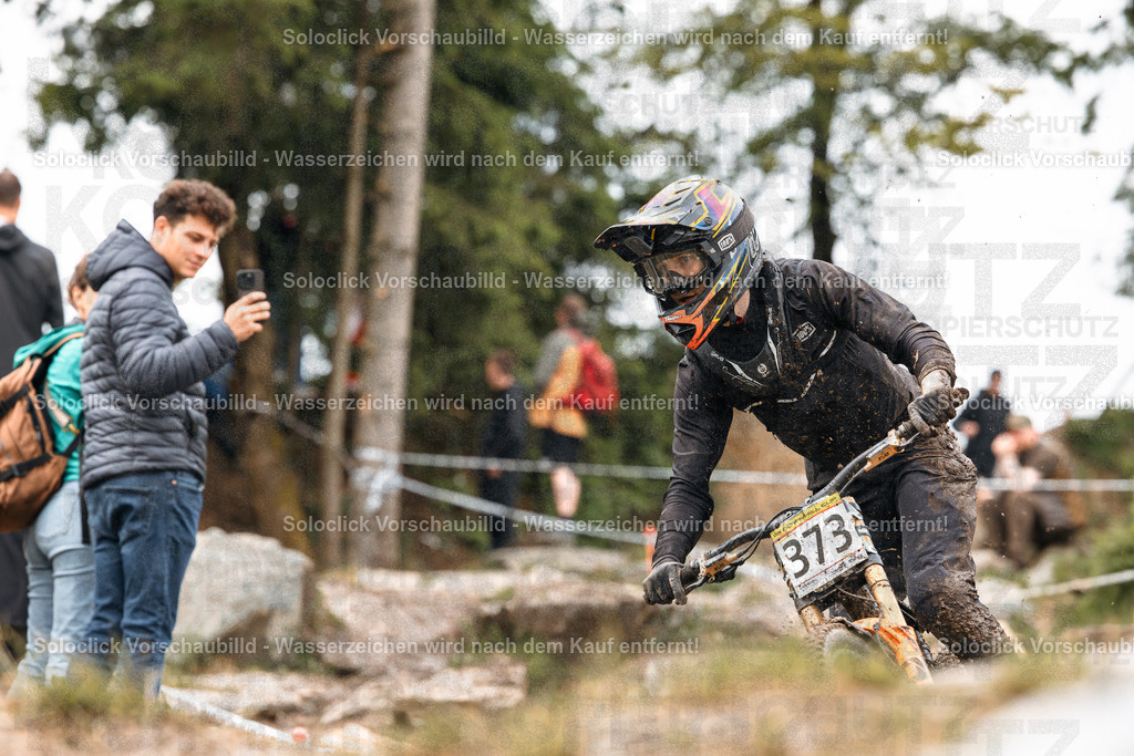 Mountainbike-Action im Wald | Ein Mountainbiker in voller Ausrüstung rast auf einem staubigen, unebenen Pfad durch einen dichten Wald. Er trägt einen schwarzen Helm mit gelben Akzenten und eine Schutzbrille. Sein Trikot ist schwarz und sein Handschuhe sind ebenfalls schwarz. Er trägt die Nummer 373 auf seinem Fahrradrahmen. Im Hintergrund sind andere Zuschauer und Bäume zu sehen. Ein Junge mit einem Rucksack hält ein Smartphone in der Hand und scheint das Rennen zu filmen. - Realisiert mit Pictrs.com