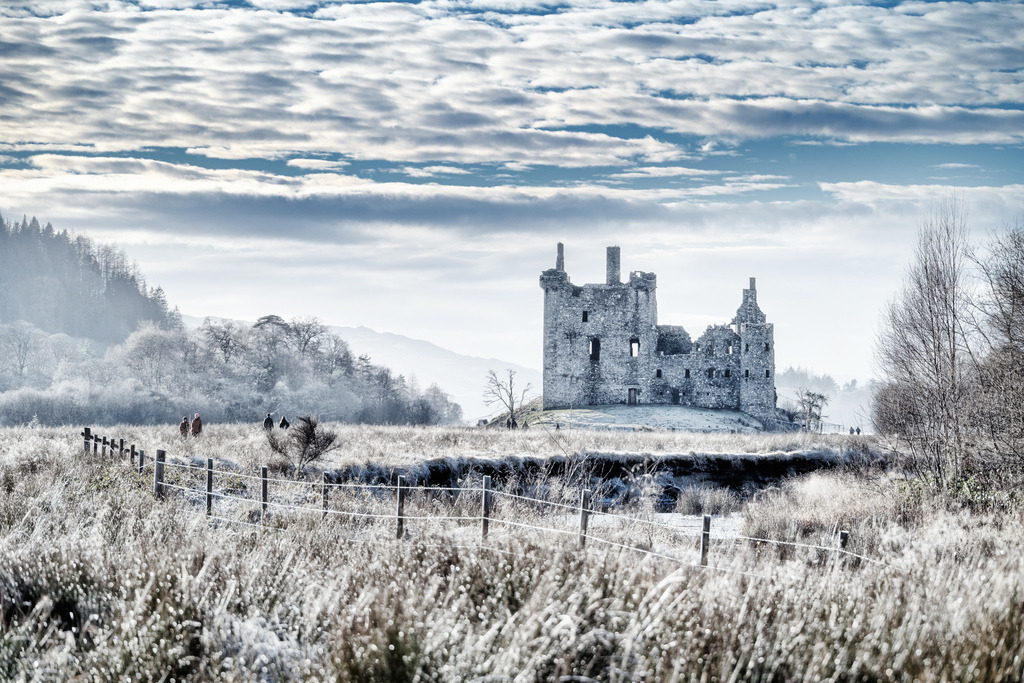 Winterlicher Frost bei Kilchurn Castle | Die Ruine von Kilchurn Castle erhebt sich majestätisch inmitten einer weiten, frostbedeckten Landschaft in Schottland. Ein niedriger Zaun im Vordergrund führt den Blick zur historischen Struktur, während der Himmel von einer dynamischen Wolkenformation geprägt wird.  - Realisiert mit Pictrs.com