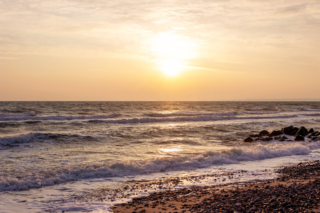 Wandbild: Sonnenaufgang an der Ostsee | Dieses Wandbild im Querformat zeigt einen schönen Sonnenaufgang an der Ostsee. Im Vordergrund ist der Sandstrand zu sehen, der vom dem Licht der aufgehenden Sonne angeleuchtet wird. Auf dem Meer sind zahlreiche Wellen. Am Himmel scheint die Sonne durch Wolken am Horizont.  - Realisiert mit Pictrs.com