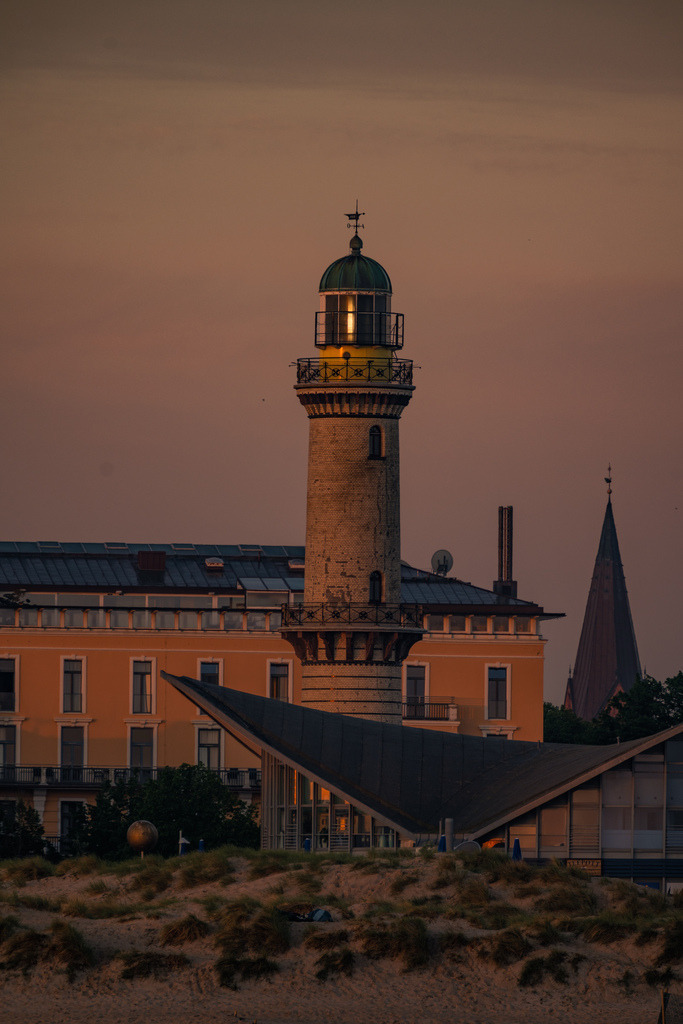 Leuchtturm Warnemünde im Abendlicht | Der historische Leuchtturm von Warnemünde erhebt sich im warmen Licht des Abends über den Gebäuden des Ostseebades. Die gedämpften Farben verleihen der maritimen Architektur eine ruhige und zeitlose Wirkung. - Realisiert mit Pictrs.com