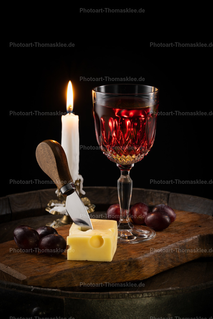 Weinprobe Arrangement: Käse, Trauben und Kerzenschein | Diese atmosphärische Weinszene präsentiert ein klassisches Rotwein-Arrangement auf rustikalem Holzbrett. Ein edles Kristallglas mit tiefrotem Wein steht neben frischem Käse mit charakteristischen Löchern und einem traditionellen Käsemesser. Dunkle Weintrauben ergänzen die Komposition perfekt, während das warme Kerzenlicht eine gemütliche Stimmung schafft. Das Holzbrett und der schwarze Hintergrund verstärken den rustikalen Charakter. Diese Szene verkörpert pure Genusskultur und lädt zum Verweilen ein.  - Realisiert mit Pictrs.com