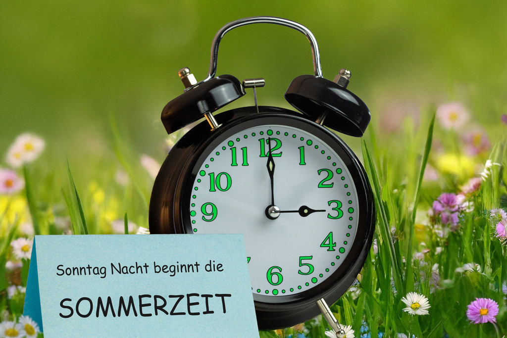 Sonntag Nacht beginnt die Sommerzeit | (Fotomontage) Zeitumstellung Sommerzeit Symbolbild: Wecker vor einer Blumenwiese und Text SONNTAG NACHT BEGINNT DIE SOMMERZEIT - Realisiert mit Pictrs.com