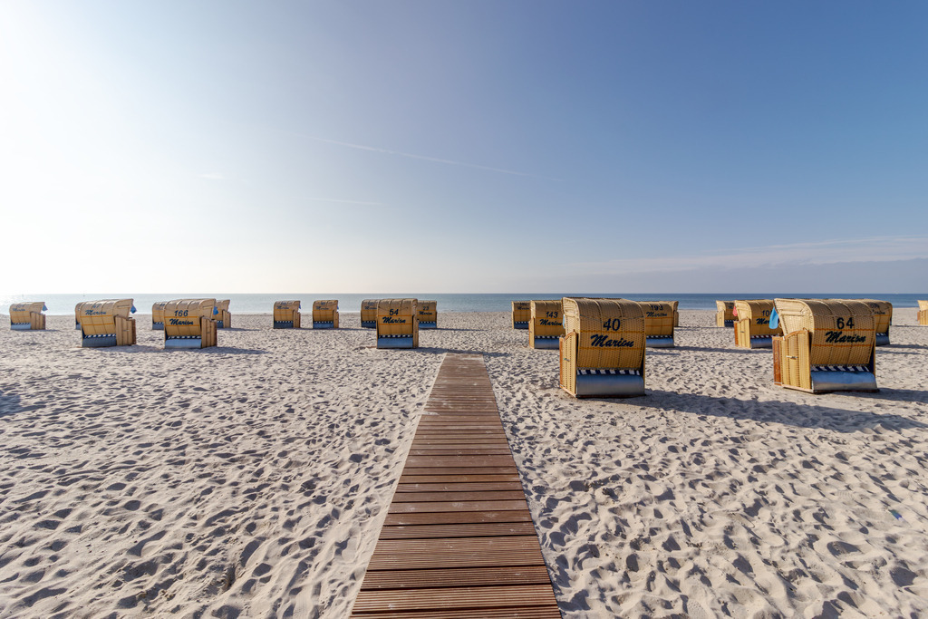 Wandbild: Strandweg mit Strandkörben – Grömitz | Dieses maritime Wandbild zeigt den Strand von Grömitz an einem sonnigen Tag. Ein hölzerner Steg führt geradlinig durch den Sand Richtung Meer und gliedert die Szene in klare Perspektiven. Links und rechts stehen zahlreiche Strandkörbe in ordentlichen Reihen und verleihen dem Bild eine strukturierte, ruhige Atmosphäre. Der Sand ist hell und fein, das Meer liegt ruhig, und der Himmel zeigt sich mit zarten Schleierwolken freundlich und weit. Die Komposition vermittelt Gelassenheit und Urlaubsstimmung – ein typisches Bild der Ostseeküste. Dieses Motiv eignet sich ideal als Wandbild für maritime Wohnkonzepte und bringt die entspannte Atmosphäre eines Sommertages direkt ins Zuhause. Erhältlich als Leinwandbild, Acrylglasbild, Alu-Dibond FineArt Print oder als Akustikbild – ein stilvoller Blickfang für Wohnzimmer, Büro oder Ferienwohnung. - Realisiert mit Pictrs.com