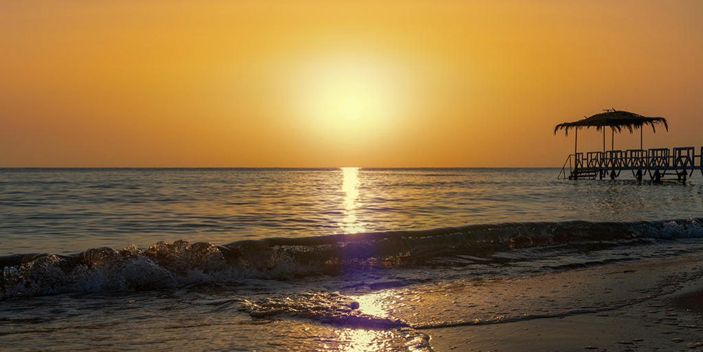 Sonnenaufgang am Roten Meer (Panorama) | Ägypten | Premium-Wandbild im Panorama-Format von justsomewhere.com: Ein goldener Sonnenaufgang über dem Roten Meer in Ägypten mit einer brechenden Welle am Strand und einem hölzernen Steg mit Strohdach als Silhouette. - Realisiert mit Pictrs.com