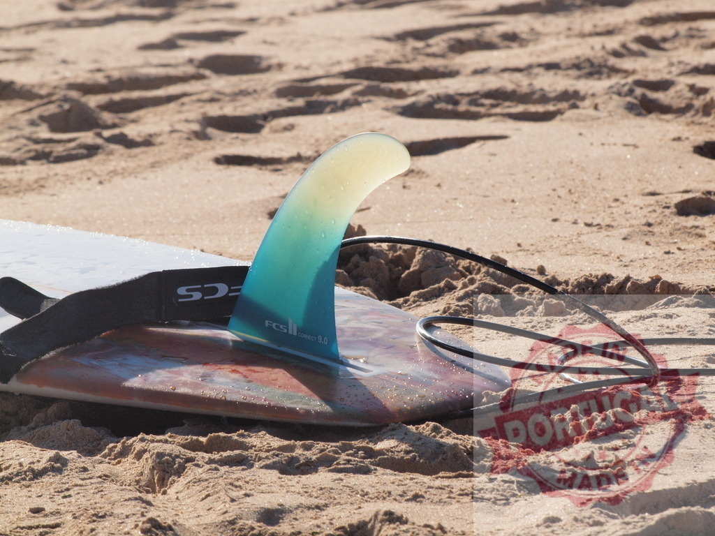 Surfboard with FCS II fin and leash resting on sandy beach | Surfboard lying on the sand with visible fin and leash, ready for surfing - Realisiert mit Pictrs.com