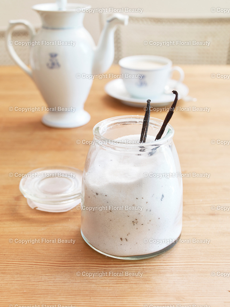 Vanillezucker | Glas gefüllt mit Zucker und Vanilleschoten auf Tisch mit Kaffeegedeck im Hintergrund. - Realized with Pictrs.com