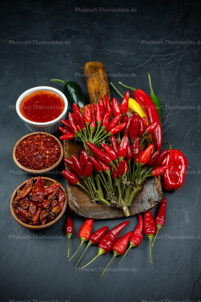 Feurige Chili-Sammlung mit Sauce und Gewürzpulver | Ein großer Strauß frischer roter Chilischoten dominiert diese appetitliche Gewürzkomposition auf dunklem Schiefer. Eine weiße Schale enthält rote Chilisauce, während zwei Holzschälchen gemahlenes Chilipulver und getrocknete Chiliflocken präsentieren. Vereinzelte grüne und gelbe Schoten ergänzen die feurige Farbpalette perfekt. Der rustikale Holzstößel unterstreicht den natürlichen Charakter dieser Küchengewürze. Diese stilvolle Anordnung zeigt die Vielfalt scharfer Gewürze und inspiriert Hobbyköche zu würzigen Kochexperimenten. - Realisiert mit Pictrs.com