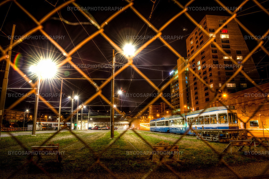 Urban Grid _ Zürich Tram at Night | Eine eindrucksvolle nächtliche Stadtszene, eingefangen hinter einem Zaun, bietet dem Betrachter einen unverfälschten und authentischen Einblick in das Zürcher Straßenbahndepot. Helle Stadtlichter, sternförmige Lichteffekte und ein Netz aus Oberleitungen erzeugen eine dynamische Komposition, während die alte Straßenbahn still im Kontrast zur pulsierenden Energie der Stadt steht.Dieser Druck besticht durch seinen starken Industriecharakter und eignet sich perfekt für moderne Interieurs, Büros und Liebhaber der Streetfotografie. - Realisiert mit Pictrs.com