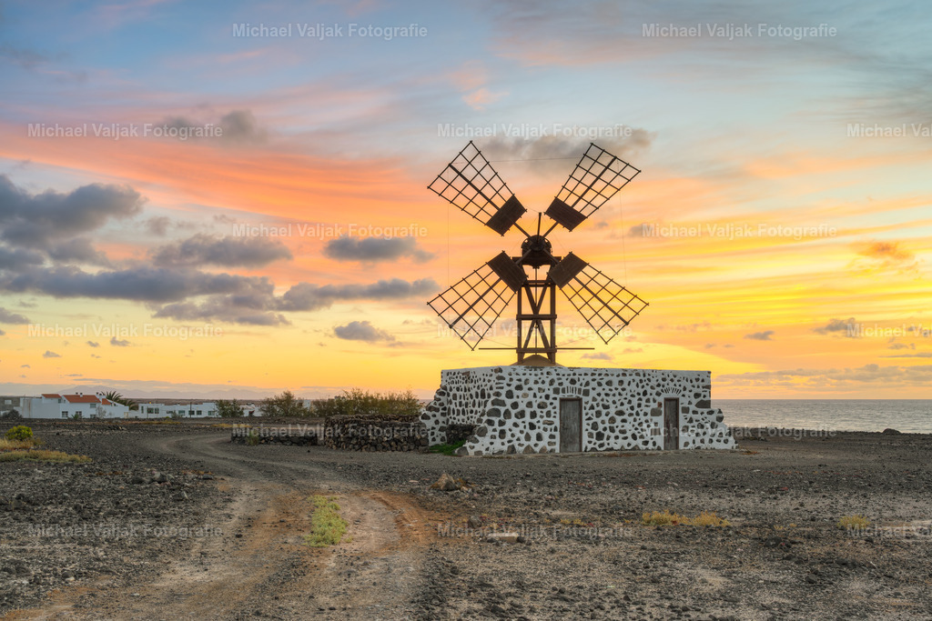 Windmühle in Puerto Lajas auf Fuerteventura | Kurz vor Sonnenaufgang steht die Windmühle von Puerto Lajas still in der kühlen Morgenluft. Das erste Licht zeichnet eine zarte Kontur um die Flügel, während der Himmel langsam von Blau zu warmen Farbtönen übergeht. Die Landschaft wirkt ruhig, fast zurückhaltend, und die Mühle steht wie ein stiller Fixpunkt in diesem sanften Übergang zwischen Nacht und Tag. - Realisiert mit Pictrs.com