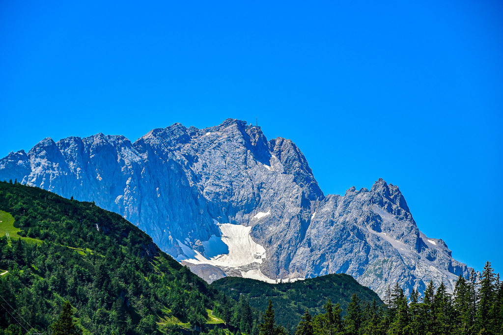 Majestät in Fels und Eis: Die Zugspitze über dem Höllental | Ein Anblick, der ehrfürchtig macht. Die mächtige Flanke der Zugspitze, Deutschlands höchstem Gipfel, ragt unbezwingbar in den strahlend blauen Himmel empor. Tief in die schroffen Felswände eingebettet, glitzert der Höllentalgletscher, ein Überbleibsel vergangener Eiszeiten und stummer Zeuge der Klimaveränderung. Die dunklen Wälder und saftig grünen Hänge im Vordergrund bilden einen markanten Kontrast zur rauen, fast unnahbaren Schönheit des Hochgebirges. Dieses Foto, festgehalten am 18. Juli 2023 bei Garmisch-Partenkirchen, ist eine Hommage an die ungezähmte Kraft und Faszination der Alpen. - Realisiert mit Pictrs.com