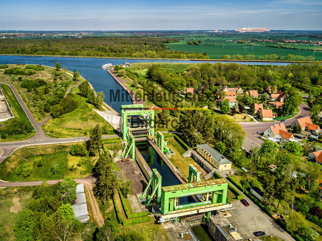 Schiffshebewerk-Rothensee_Luftbild_Magdeburg_technisches_Denkmal-0048 | Das Luftbild zeigt das Schiffshebewerk Rothensee aus geringer Höhe an einem sonnigen Sommertag. Die markante Stahlkonstruktion erhebt sich eindrucksvoll über die umgebende Wasserlandschaft und vermittelt aus der Drohnenperspektive einen hervorragenden Eindruck von den Dimensionen dieses außergewöhnlichen Ingenieurbauwerks. Die Nähe der Aufnahme lässt die filigranen Fachwerkkonstruktionen und die technischen Details besonders gut erkennen.Das Schiffshebewerk Rothensee wurde zwischen 1934 und 1938 errichtet und nahm 1938 den Betrieb auf. Mit einer Hubhöhe von 16 Metern verbindet es den Mittellandkanal mit dem Elbe-Havel-Kanal. Der wassergefüllte Trog ist 85 Meter lang und 12 Meter breit und kann Schiffe mit einem Gesamtgewicht von bis zu 5.400 Tonnen anheben oder absenken. Das Hebewerk arbeitet nach dem Prinzip des Gewichtsausgleichs, wodurch für einen Hebevorgang vergleichsweise wenig Energie benötigt wird.Die Drohnenaufnahme verdeutlicht eindrucksvoll die technische Raffinesse und die außergewöhnliche Konstruktion des Schiffshebewerks. Als eines der bedeutendsten Wasserbauwerke Deutschlands steht es bis heute für innovative Ingenieurkunst und spielt gemeinsam mit der benachbarten Schleuse eine wichtige Rolle im europäischen Binnenwasserstraßennetz. - Realisiert mit Pictrs.com