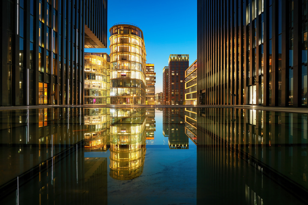 Wandbild Nächtliche Eleganz: Glanz der modernen Architektur | Dieses atemberaubende Wandbild zeigt eine moderne Stadtszene bei Nacht, in der sich die eindrucksvollen Glasfassaden der Gebäude in einem ruhigen Wasserbecken spiegeln. Die beleuchteten Gebäude leuchten in warmen Gelb- und Orangetönen, die im Kontrast zu dem tiefblauen Nachthimmel stehen. Die Symmetrie der Architektur und die perfekte Spiegelung im Wasser schaffen ein Gefühl von Ruhe und Harmonie. Dieses Bild fängt die Schönheit und den Glanz urbaner Landschaften ein und lädt den Betrachter ein, die moderne Eleganz und die Ruhe der Nacht zu genießen.