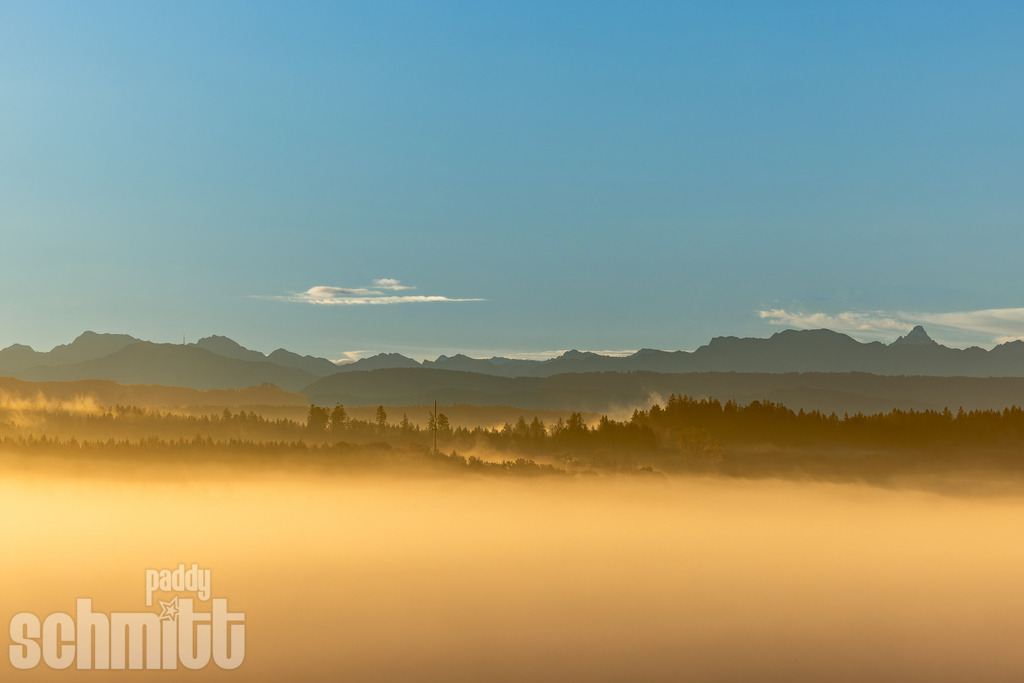 Warmer Morgennebel mit Bergpanorama im Allgäu | Warmer Morgennebel liegt über den Tälern des Allgäus und taucht die Landschaft in ein sanftes, goldenes Licht. Im Hintergrund erhebt sich das beeindruckende Bergpanorama – eine stille und stimmungsvolle Momentaufnahme des frühen Morgens. - Realisiert mit Pictrs.com