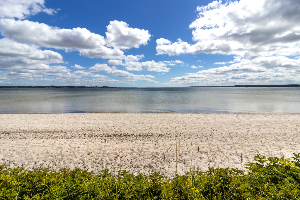 Wandbild: Strand in Holnis im Sommer | Dieses Wandbild im Querformat zeigt den Strand in Holnis im Sommer. Am blauen Himmel sind zahlreiche Wolken zu sehen die sich auf dem Wasser der Flensburger Förde spiegeln. - Realisiert mit Pictrs.com