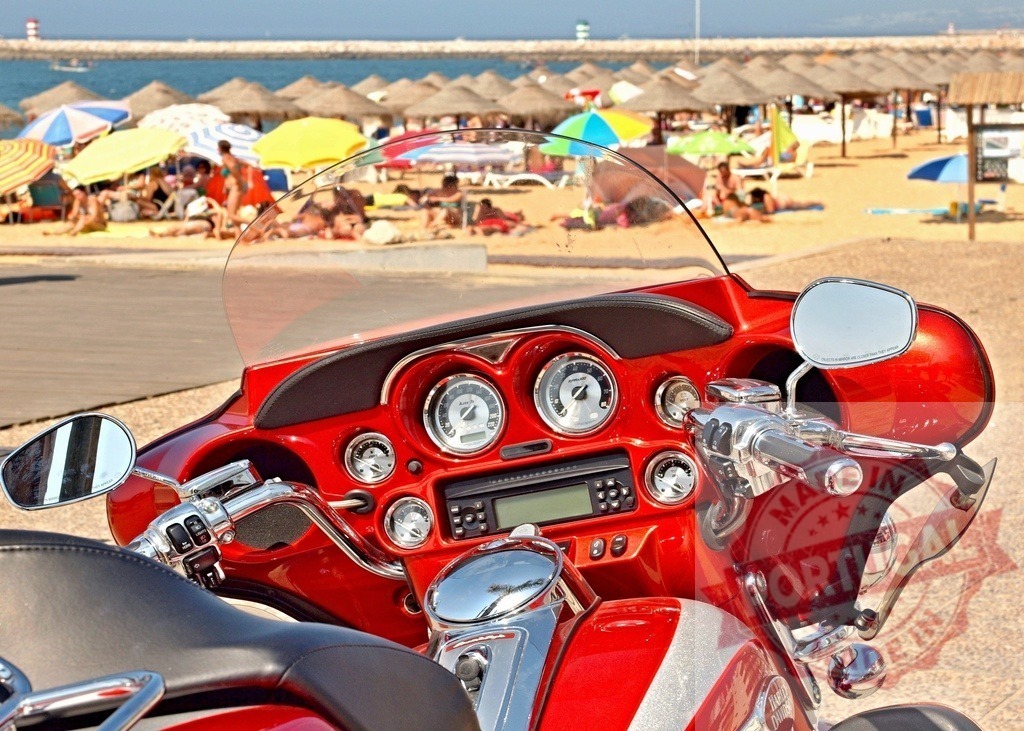 Red touring motorcycle parked at sunny beach resort | Red motorcycle dashboard and handlebars with a busy beach in the background - Realized with Pictrs.com