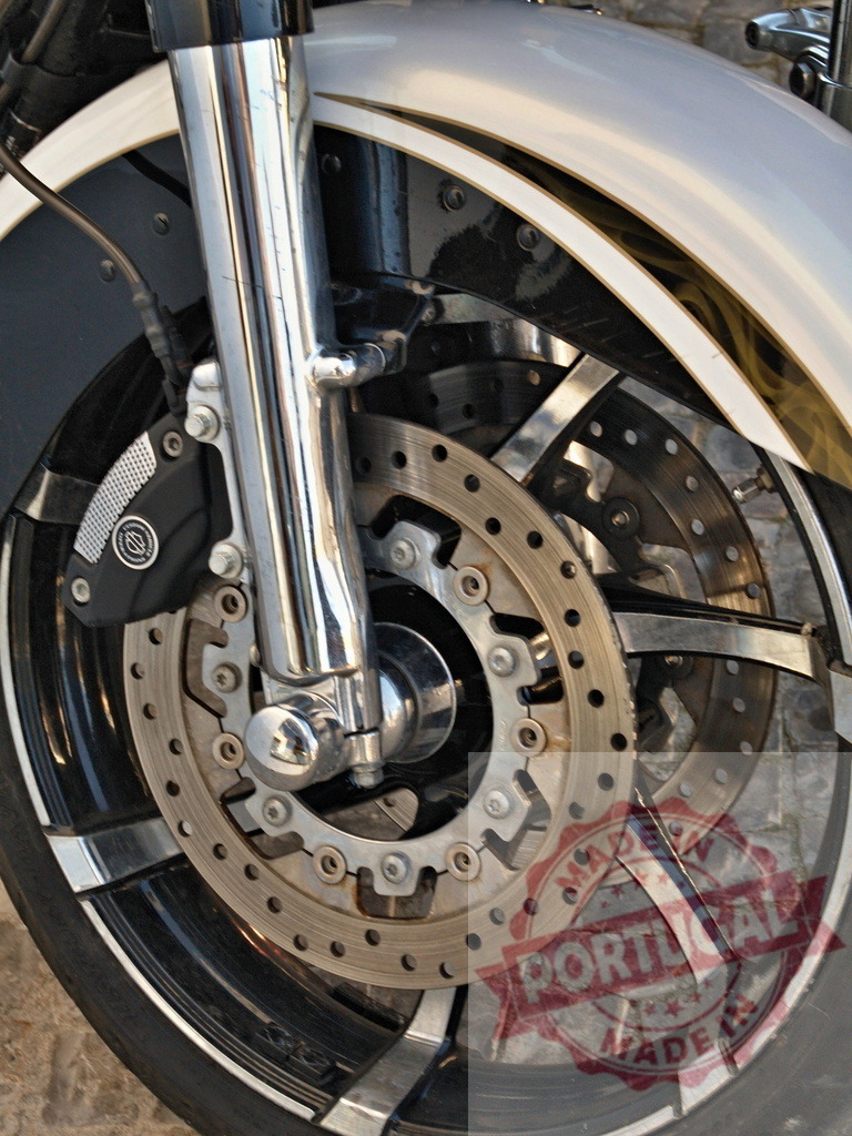 Harley-Davidson motorcycle front wheel showing chrome and disc brakes | Motorcycle front wheel with shiny chrome components, dual disc brakes, and tire on cobblestone - Realized with Pictrs.com