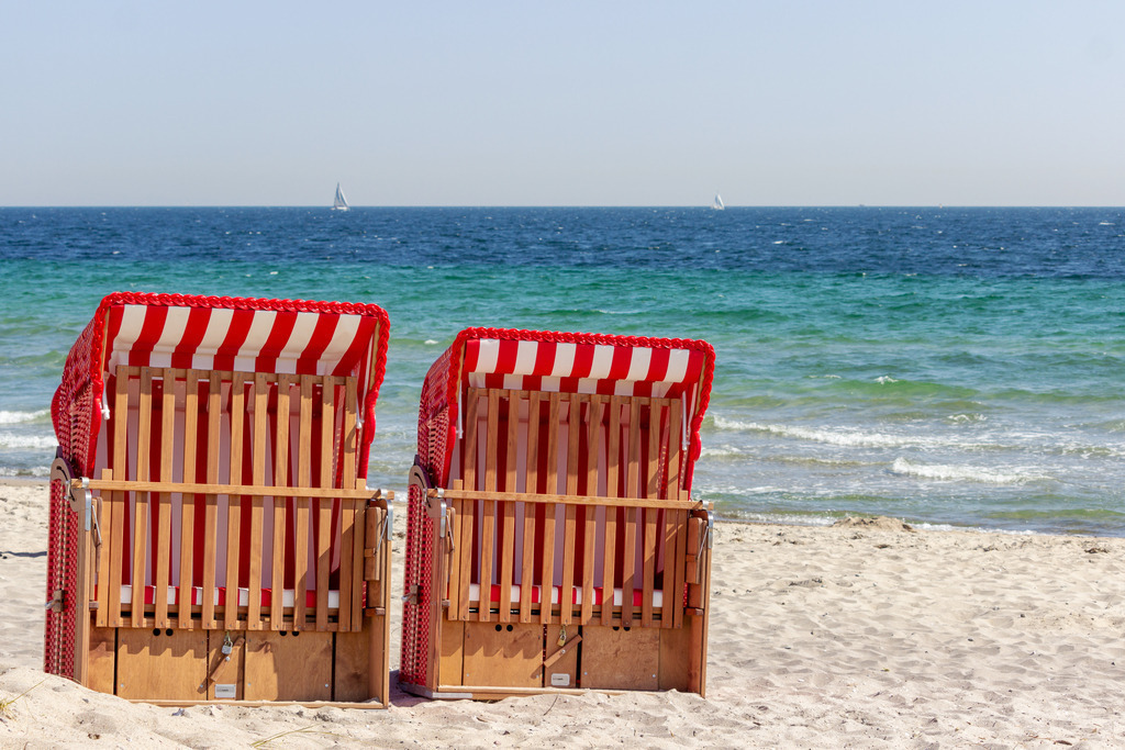 Wandbild: Strandkörbe am Strand in Schönhagen | Zwei rote Strandkörbe stehen auf dem feinen Sandstrand von Schönhagen, leicht versetzt nebeneinander. Im Hintergrund gleiten Segelboote gemächlich über die ruhige Ostsee, deren Oberfläche in zarten Blau- und Grüntönen schimmert. Dieses Wandbild verbindet sommerliche Leichtigkeit mit maritimer Klarheit – ein stiller Moment an der Küste, der zum Innehalten einlädt. - Realisiert mit Pictrs.com