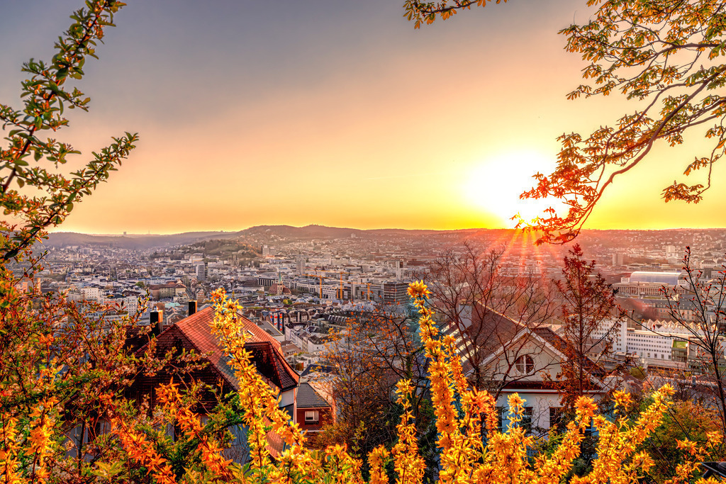 Sonnenaufgang über die sanften Hügel von Stuttgart | Das Bild zeigt einen malerischen Sonnenaufgang über Stuttgart, aufgenommen möglicherweise von der Uhlandshöhe. Es fängt die Stadtlandschaft im warmen Licht der aufgehenden Sonne ein. Die Perspektive bietet einen weiten Blick über das Stadtzentrum und die umliegenden Hügel.Im Vordergrund sind herbstlich gefärbte Büsche und Dächer von Wohnhäusern zu sehen. - Realisiert mit Pictrs.com