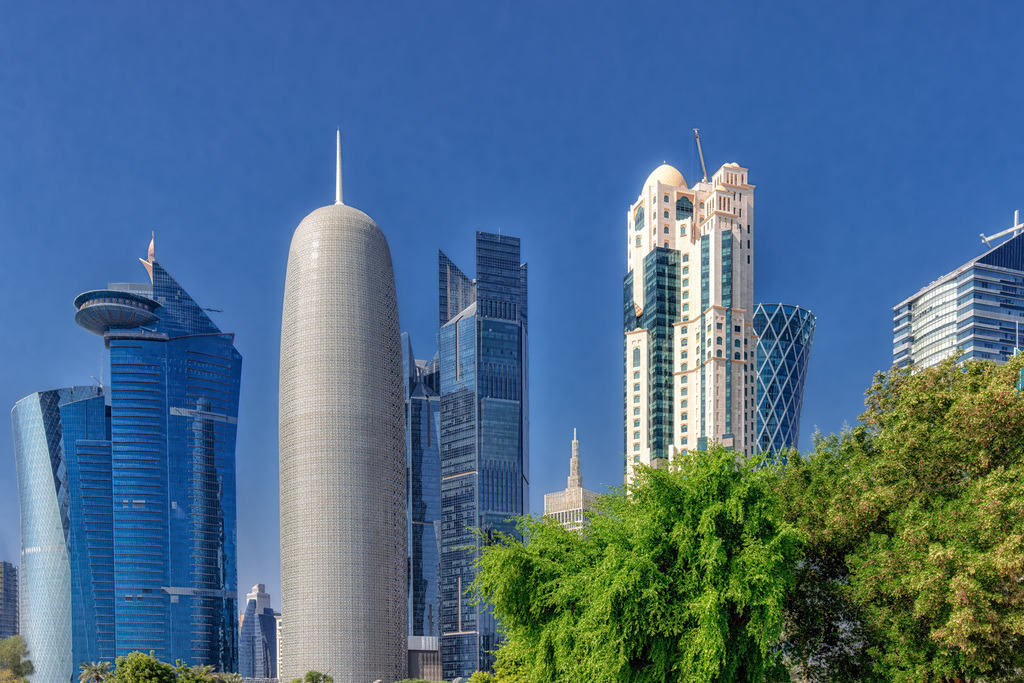Skyline der Westbay in Doha,Mashrabiya-Ornamente,Palm Tower | Das Bild zeigt die markante Skyline des Geschäftsviertels West Bay in Doha, Katar, entlang der Küstenstraße Doha Corniche.Doha Tower (Burj Doha): Der auffällige, zylinderförmige und kuppelförmige Wolkenkratzer im Vordergrund (entworfen von Jean Nouvel) prägt das Stadtbild mit seiner an arabische Mashrabiya-Ornamente erinnernden Fassade.Architekturmix: Links daneben ist das markante Hochhaus mit der auskragenden Aussichtsplattform (Palm Tower oder angrenzende Bauwerke) zu sehen, während rechts weitere moderne Hochhäuser wie der World Trade Center Doha oder der Tornado Tower das Finanzzentrum säumen. - Realisiert mit Pictrs.com
