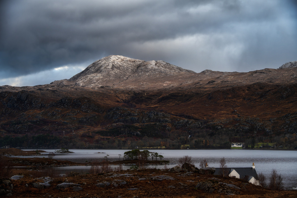 Loch Maree | Eine weite Ansicht des Loch Maree in Schottland zeigt schneebedeckte Berggipfel unter einem dramatischen Wolkenhimmel. Im Vordergrund erstreckt sich der See mit kleinen Inseln und einem traditionellen Haus am Ufer, umgeben von karger, winterlicher Vegetation. Die Komposition betont die raue Schönheit der schottischen Highlands und die Weite der unberührten Natur. - Realisiert mit Pictrs.com