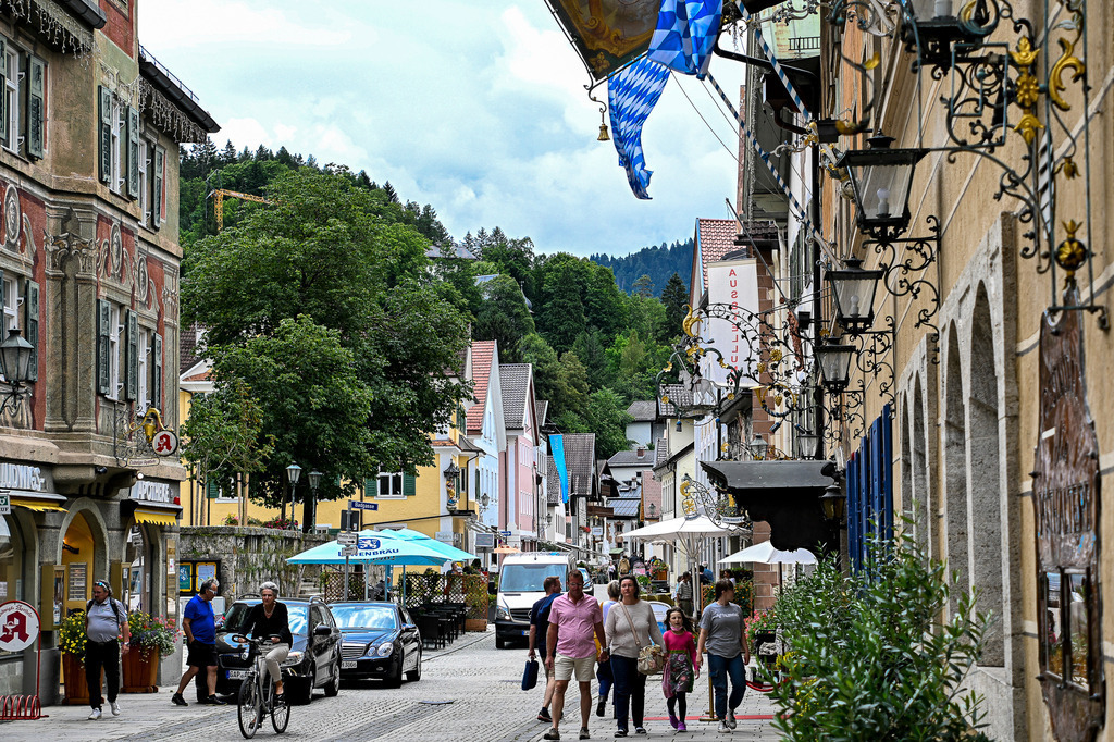 Traditioneller Flair und Sommerleben in Partenkirchen | Prachtvolle, historisch bemaltes Gemäuer, schwebende Rautenfahnen und verzierte Laternen rahmt das bunte Sommertreiben in der Partenkirchener Ludwigstraße ein. Während Spaziergänger entspannt durch die verkehrsberuhigte Gasse schlendern, verbinden sich aufwendige Lüftlmalereien und traditionelle Baukunst mit alpenländischem Lebensgefühl. Die Kulisse strahlt eine wohltuende Entschleunigung aus und lädt ein, die reiche Kulturgeschichte Partenkirchen im Schatten der nahen Bergwälder hautnah zu genießen. - Realisiert mit Pictrs.com