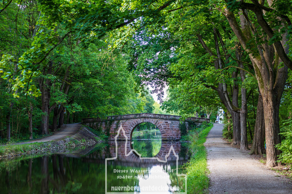 Monatsbild des Kalender 2026 für den Monat September | Eine historische Steinbogenbrücke überspannt den Ludwig-Donau-Main-Kanal in Nürnberg, Bayern. Die ehemalige Schifffahrtsstraße ist von dichtem Baumbestand gesäumt und dient heute als Naherholungsgebiet inmitten des Stadtgebiets. Die ruhige Wasseroberfläche spiegelt Brücke und Umgebung. - Realisiert mit Pictrs.com