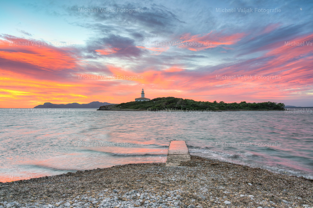 Blick zum Leuchtturm von Alcanada auf Mallorca | Kurz vor Sonnenaufgang liegt die Küste Mallorcas noch in sanftem Dämmerlicht, während der Himmel bereits in kräftigen Rot‑ und Orangetönen leuchtet. Die Insel von Alcanada ist deutlich erkennbar: der Leuchtturm, die Felsen, die niedrige Vegetation – alles zeigt sich in weichem, natürlichem Licht, das noch keine harten Schatten wirft. Das Meer im Vordergrund wirkt ruhig und trägt nur einen feinen Schimmer der warmen Farben, wodurch der Blick automatisch zur Insel geführt wird. - Realisiert mit Pictrs.com