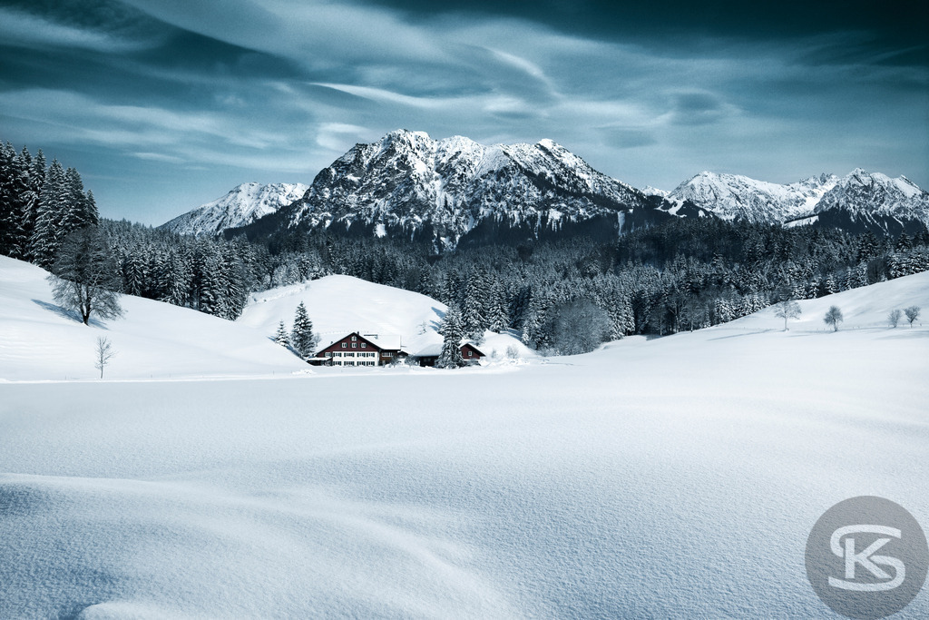 Idylle in den winterlichen Alpen: Traditionelles Haus vor Bergpanorama | Eine idyllische Szene mit einem traditionellen, schneebedeckten Alpenhaus, eingebettet in eine weite, unberührte Schneelandschaft, umgeben von einem dichten Nadelwald und überragt von einem markanten, schneebedeckten Bergmassiv im Hintergrund. - Realisiert mit Pictrs.com