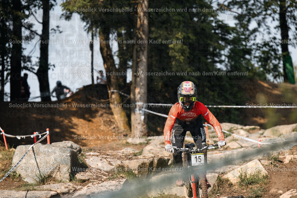 Mountainbiker im Rennen | Ein Mountainbiker in voller Ausrüstung rast durch einen staubigen, bewaldeten Kurs. Er trägt einen roten Helm und einen roten Suit mit dem Logo von Fox Racing. Er fährt ein Mountainbike mit breiten Reifen und einer Federgabel. Der Kurs ist mit Steinen und Wurzeln übersät und wird von Bäumen und Büschen gesäumt. Im Hintergrund sind Zuschauer zu sehen. - Realisiert mit Pictrs.com