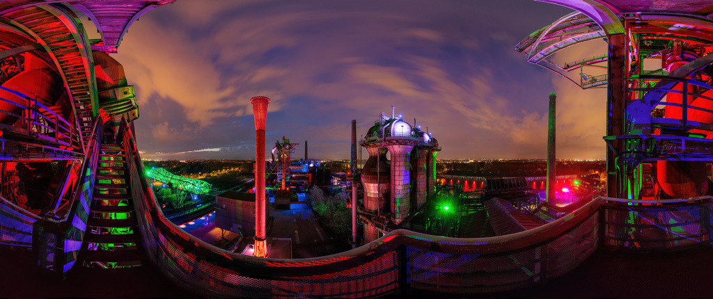 Wandbild farbenfrohe Industriearchitektur bei Nacht | Dieses atemberaubende Wandbild zeigt eine industrielle Anlage bei Nacht, die durch leuchtende und farbenfrohe Beleuchtung hervorgehoben wird. Im Vordergrund befinden sich eine metallene Treppe und verschiedene industrielle Strukturen, die in intensiven Rot-, Grün- und Blautönen erstrahlen. Die Mitte des Bildes wird von hohen Schornsteinen und monumentalen Gebäuden dominiert, die sich gegen den dramatisch beleuchteten Nachthimmel abzeichnen. Der Himmel selbst ist mit dynamischen Wolkenformationen bedeckt, die von den Lichtern der Stadt erleuchtet werden und eine fesselnde und fast futuristische Stimmung erzeugen. Die Weite des Industriegeländes erstreckt sich bis zum Horizont, wo die Lichter der Stadt in der Ferne glitzern.