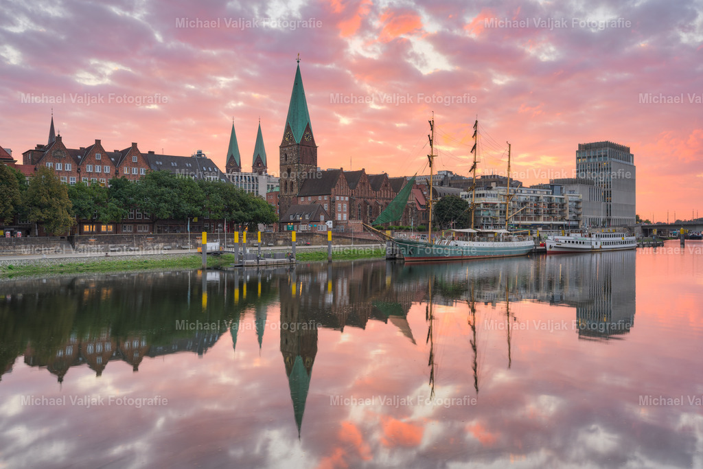 Blick zur Schlachte in Bremen bei Sonnenaufgang | Ein Blick über die Weser in den frühen Morgenstunden zeigt die Altstadt im warmen Licht eines klaren Sonnenaufgangs. Die Fassaden der historischen Gebäude spiegeln sich deutlich im ruhigen Wasser und verleihen der Szene eine besondere Atmosphäre. Das Segelschiff Alexander von Humboldt liegt gut sichtbar im Vordergrund und ergänzt das Stadtbild mit seinem markanten Erscheinungsbild. Die Kombination aus Fluss, Architektur und Schiff vermittelt ein harmonisches Bild der Stadt am Morgen. - Realisiert mit Pictrs.com
